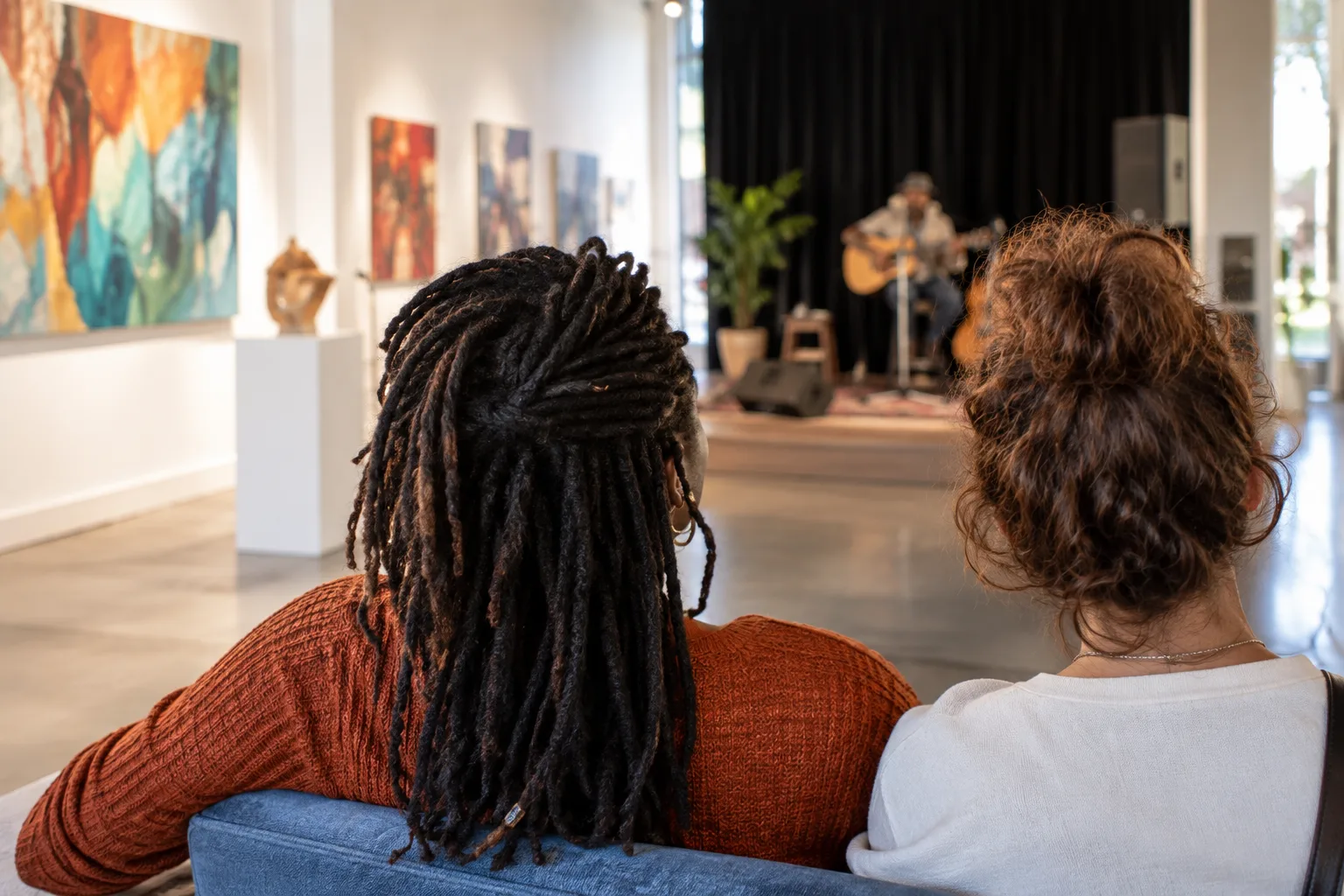 A bright local gallery beside a community theater, with artwork on display and a small live performance underway.